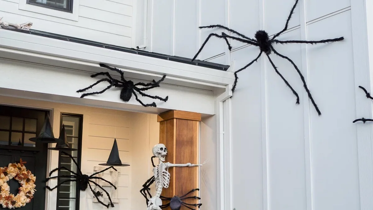 Modern black and white exterior porch featuring oversized black spiders climbing siding, a climbing skeleton, and buffalo check pillows