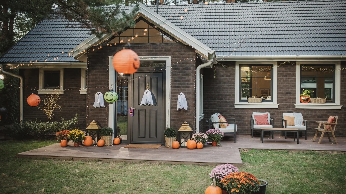 Charming dark wood cottage porch decorated with stacked cream pumpkins, warm string lights, and delicate hanging fabric spirits